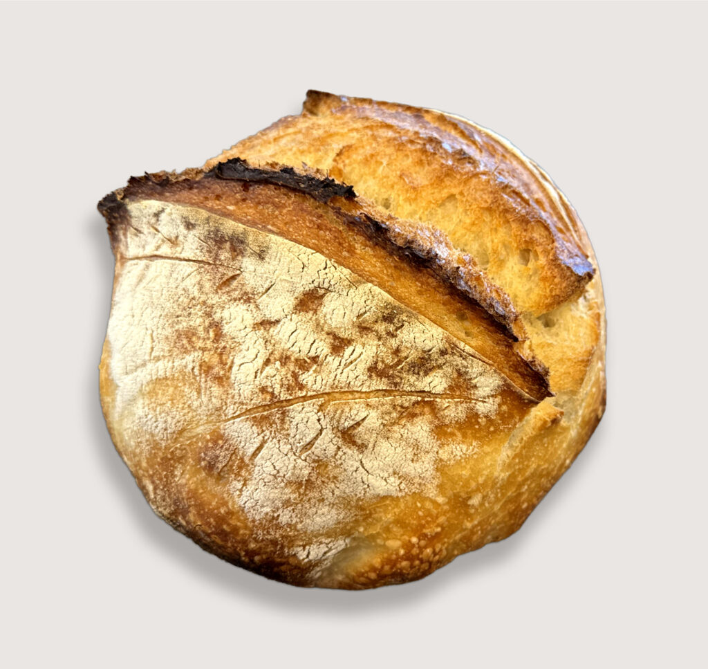 Round loaf of rustic sourdough bread with a browned, crusted exterior and a split top revealing the baked interior, lightly dusted with flour.